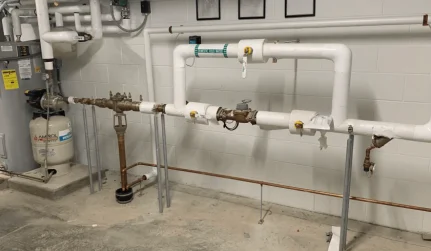 Copper water service riser with brass shutoff valves and a backflow assembly mounted on a white basement wall, PVC drain along the floor, Jackson Plumbing