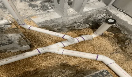 White PVC drain lines with purple primer marks laid in a gravel trench under a slab, a wye joint in the foreground, Jackson Plumbing